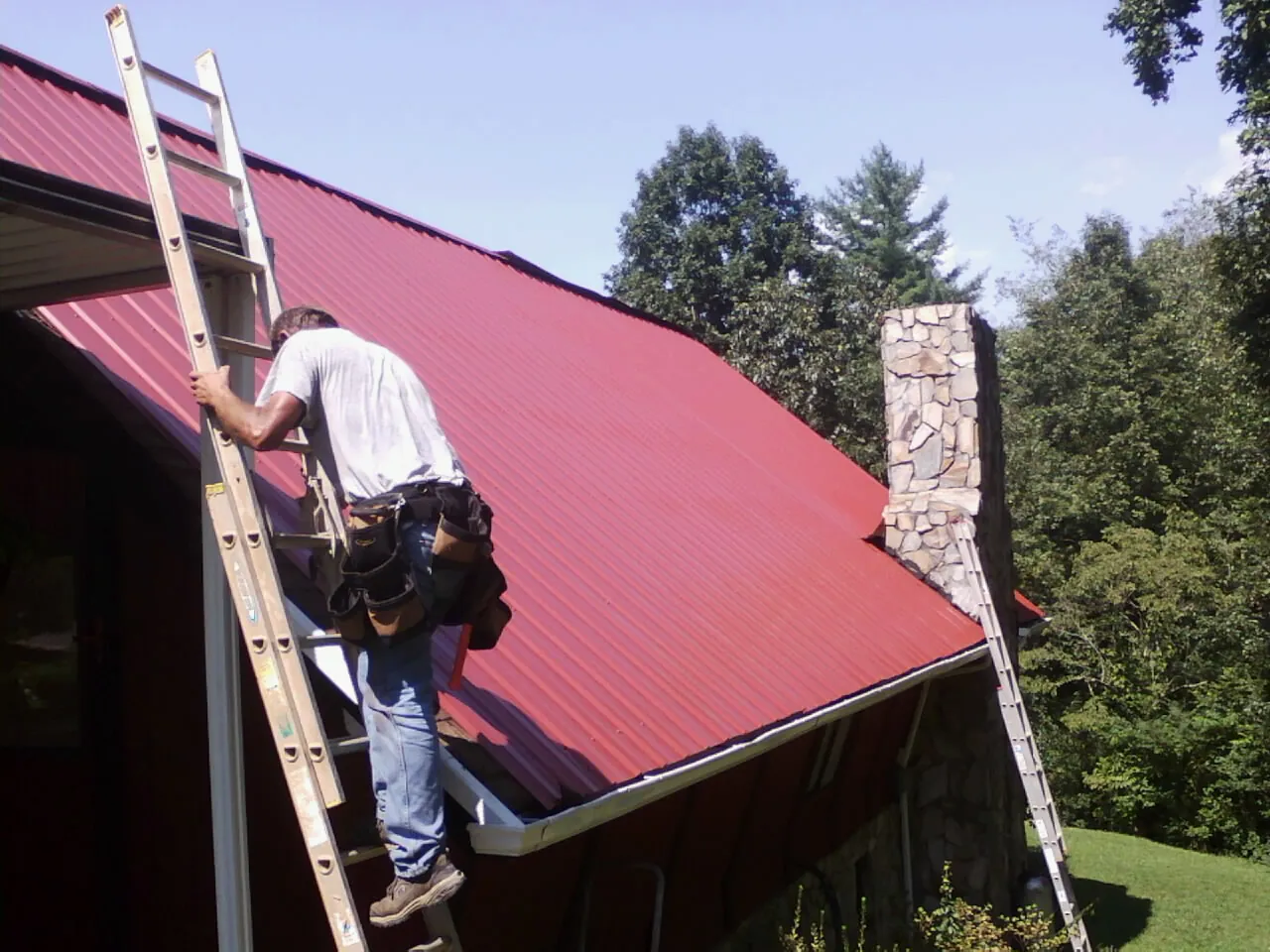 Roofer on a ladder beside a red metal roof and stone chimney — Soffit & Fascia Repair by Roofing Brothers in South Pasadena